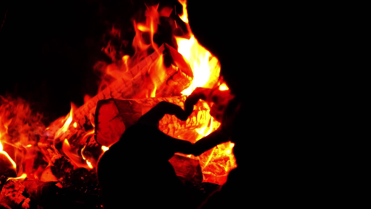A woman makes the heart sign in a forest at night by a fire. The heart is in front of the fire and is illuminated. Slow motion