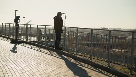 a father carrying his son walks towards a metal railing, pausing to gaze at the cityscape below, both are dressed in jackets and beanies, with the sun casting a long shadow on the pavement