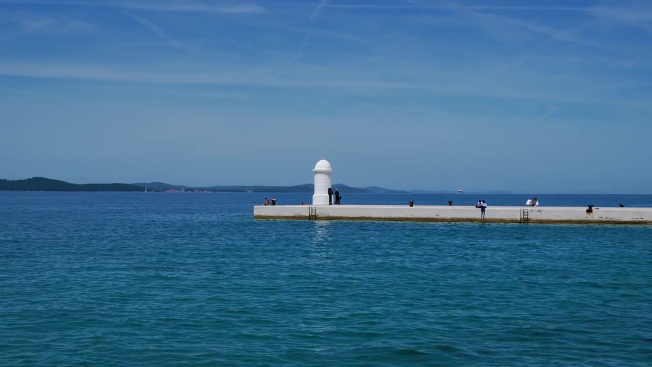 Persone che camminano lungo il molo di pietra bianca con il faro della Riva di Zara attraverso lo scintillante mare blu.
