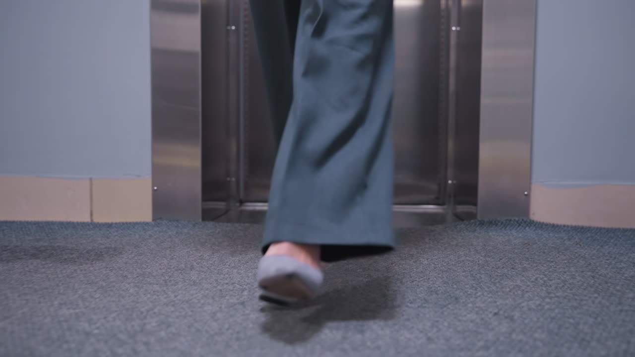 Close-up shot of businesswoman's feet stepping out of an elevator in gray shoes, wearing wide pants, transitioning into modern office environment with sleek metallic elevator interior