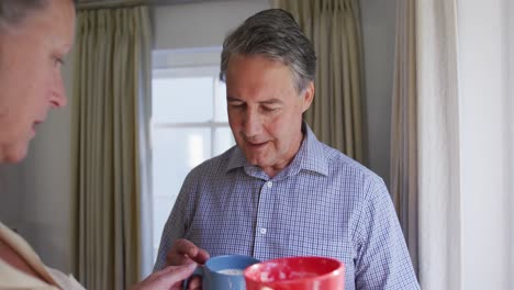 Happy-senior-caucasian-couple-looking-out-of-window-in-living-room,-drinking-coffee-and-talking