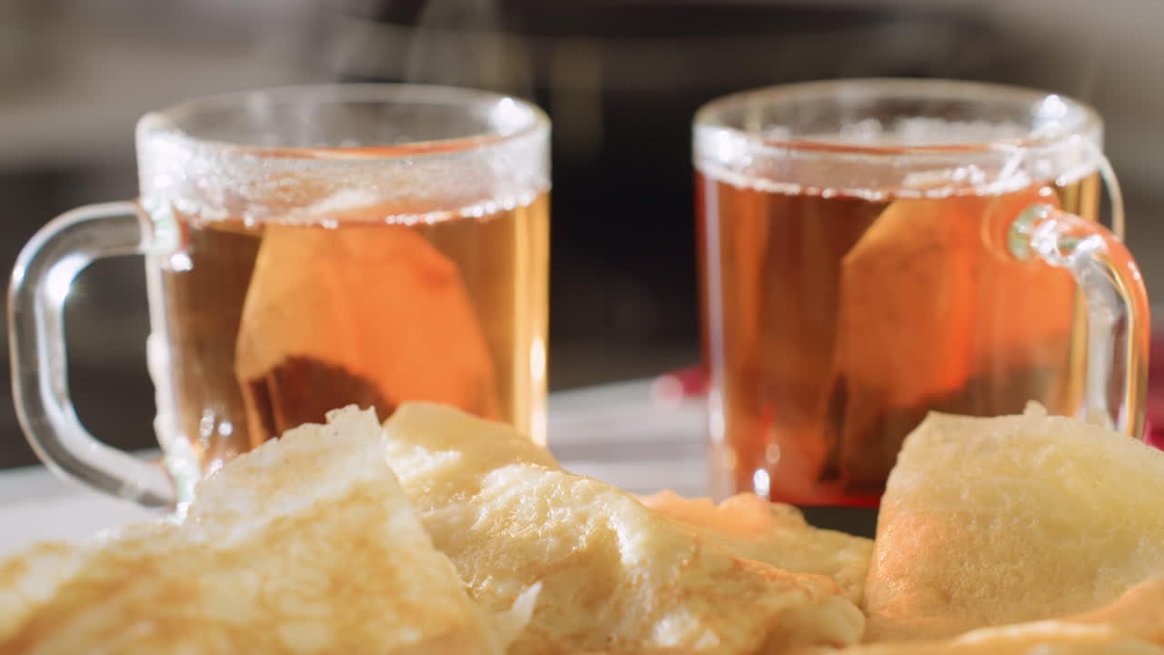 Close up of golden pancakes resting on plate with two steaming cups of hot tea in background, warm morning light highlighting fresh breakfast meal
