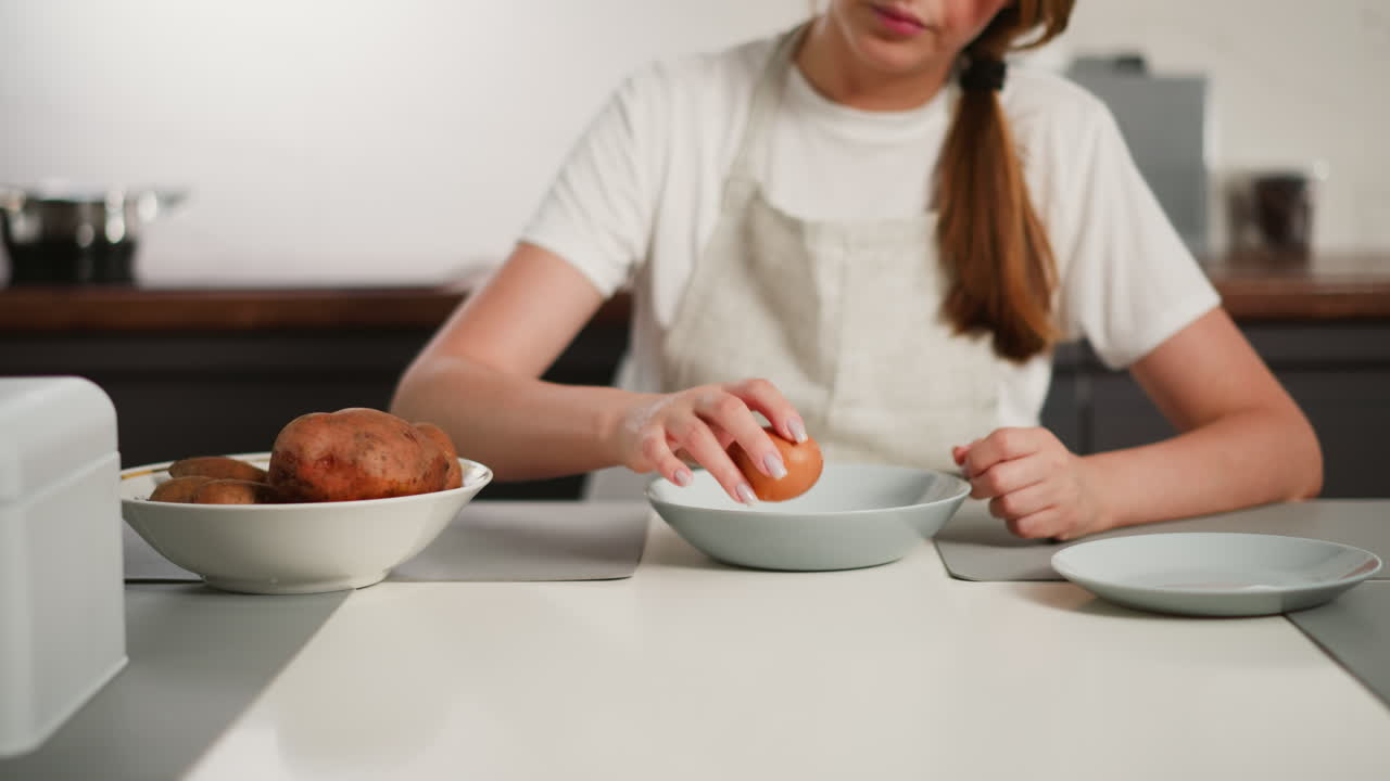 Close up of female chef wearing apron holding egg over glass plate, preparing to crack it while seated at table with raw potatoes and dishes in clean modern kitchen during food preparation routine
