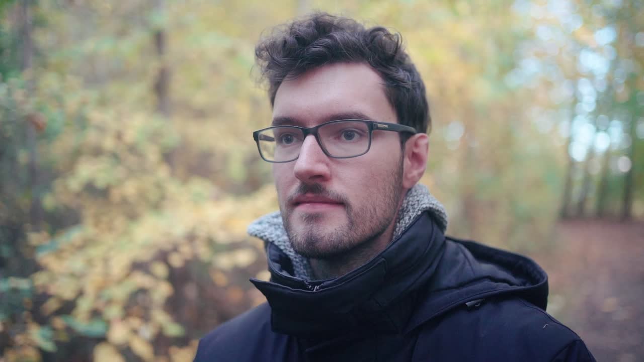 en cautivadora cámara lenta, un joven europeo con gafas afiladas y una barba elegante encuentra consuelo en un bosque mixto de otoño, su mirada atravesando el lejano horizonte, perdido en sus pensamientos