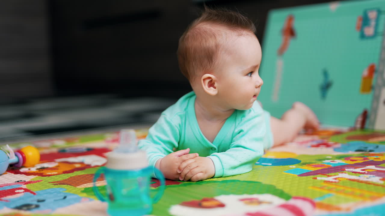 Playful sweet kid lies on belly waving feet. Tiny child stretches arm to take a bottle. Beautiful baby sticking out his tongue.