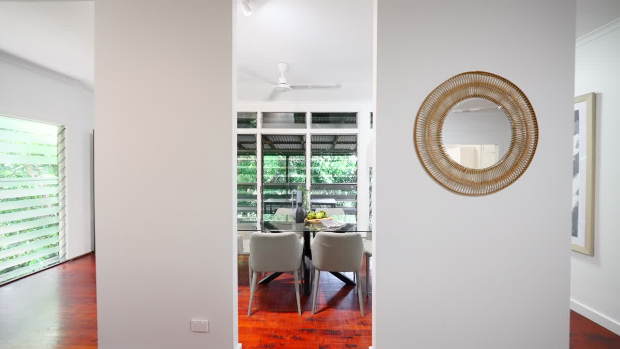 Carrello verso l'interno di una casa moderna con pavimento in legno, zona pranzo rialzata con tavolo in vetro e sedie grigie, ciotola di frutta decorativa sotto finestre a gelosia.