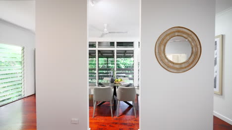 Dolly-to-Modern-Home-Interior-Wooden-Flooring-Elevated-Property-Dining-Area-with-Glass-Table-and-Grey-Chairs,-Staged-Fruit-Bowl-Below-Jealousie-Windows