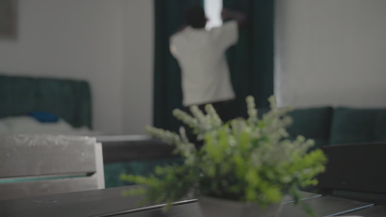 Person in white t-shirt walking through minimalist home, passing a table with small plant, sunlight streaming through dark green curtains, and modern furniture visible in the background