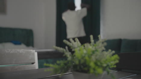 person in white t-shirt walking through minimalist home, passing a table with small plant, sunlight streaming through dark green curtains, and modern furniture visible in the background