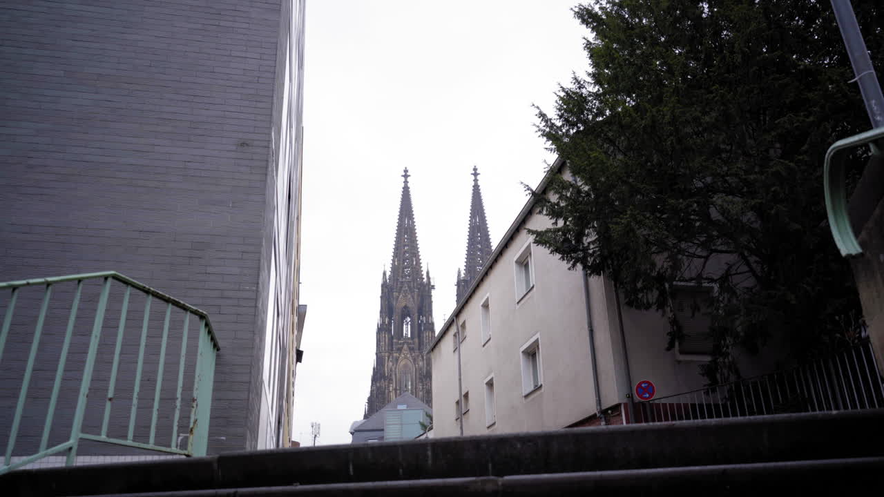 A unique perspective of Cologne Cathedral's iconic spires framed by modern urban surroundings, viewed from the top of a stairway on a cloudy day.