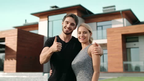 una pareja feliz haciendo un gesto de pulgares hacia arriba cerca de una casa de lujo.