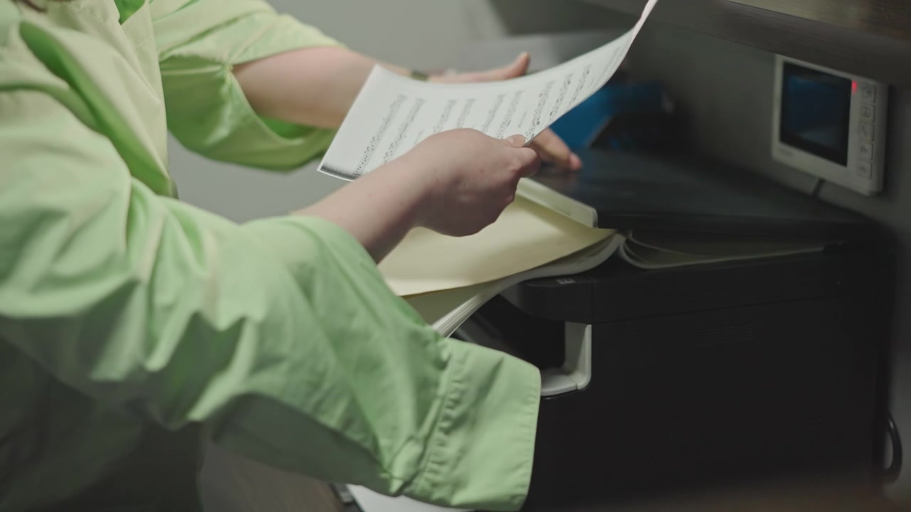 Caucasian music teacher copying sheet music from printer, hands sorting pages in green jacket, organizing stacked scores for school choir rehearsal, closeup on fingers flipping notation, calm focused