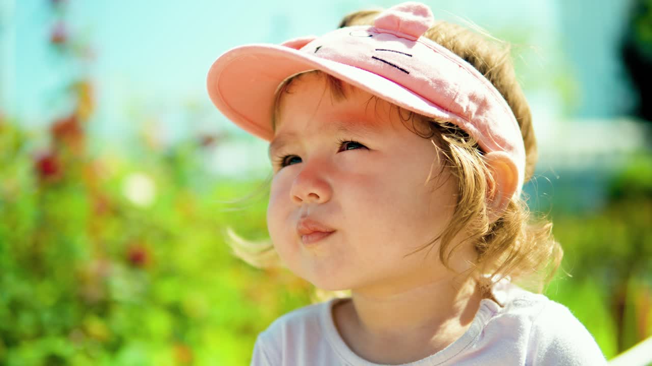 ピンクの帽子をかぶって、夏の晴れた日に庭でよそ見をしている小さな金髪の幼児の女の子の夢を見る顔