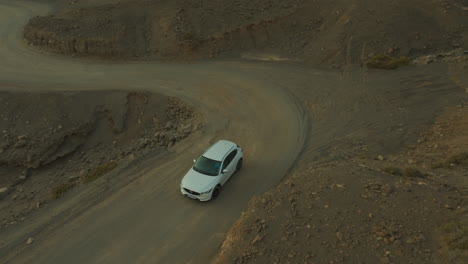 Fantastic-aerial-view-with-tracking-of-a-white-car-driving-along-a-winding-dirt-road-in-a-desert-and-volcanic-environment