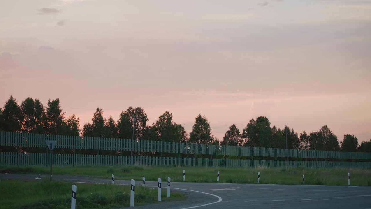 curved country road lined with white posts and green grass bordered by tall trees under pink sunset sky with rustic fence creating serene park vibe at twilight on open landscape