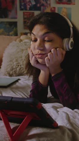 african american girl looks at tablet display while watches movie or video content. teenage girl in headphones lies on bed in her room and uses tablet computer. vertical shot