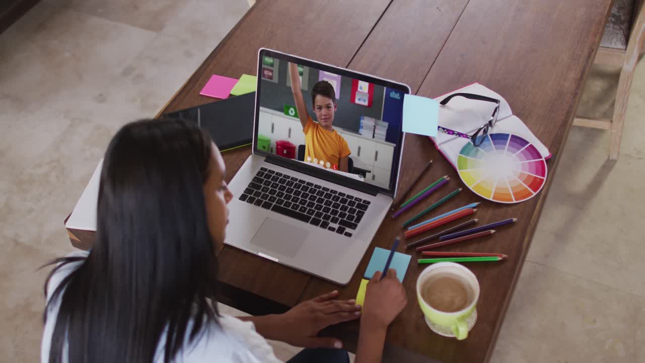 Maestra afroamericana escribiendo en notas adhesivas mientras realiza una videollamada con un niño en su computadora portátil.