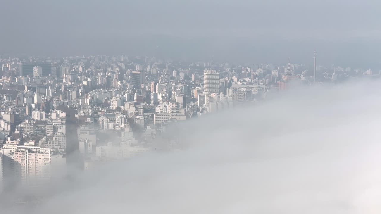 Aerial drone smoothly navigates Montevideo as thick fog advances, wrapping towers and streets in a soft white veil
