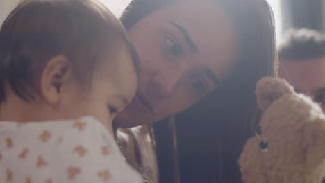 Close-Up-Of-A-Happy-Mother-In-Bed-Holding-A-Teddy-Bear-And-Playing-With-Her-Little-Daughter-While-Dad-Watching-Them