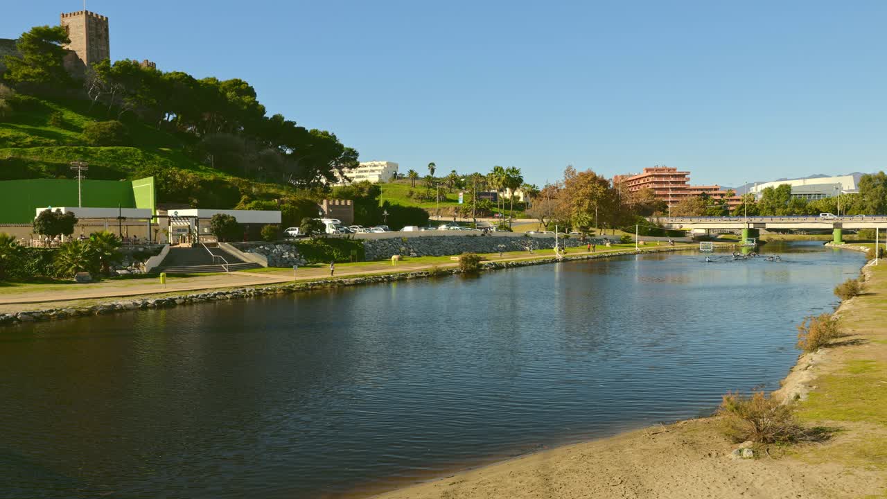 Ampia panoramica a sinistra di un fiume a Fuengirola, Málaga, Costa del Sol, Spagna, con giocatori di canoa polo sull'acqua e pedoni che passeggiano lungo la sponda soleggiata, mostrando una vivace attività all'aperto