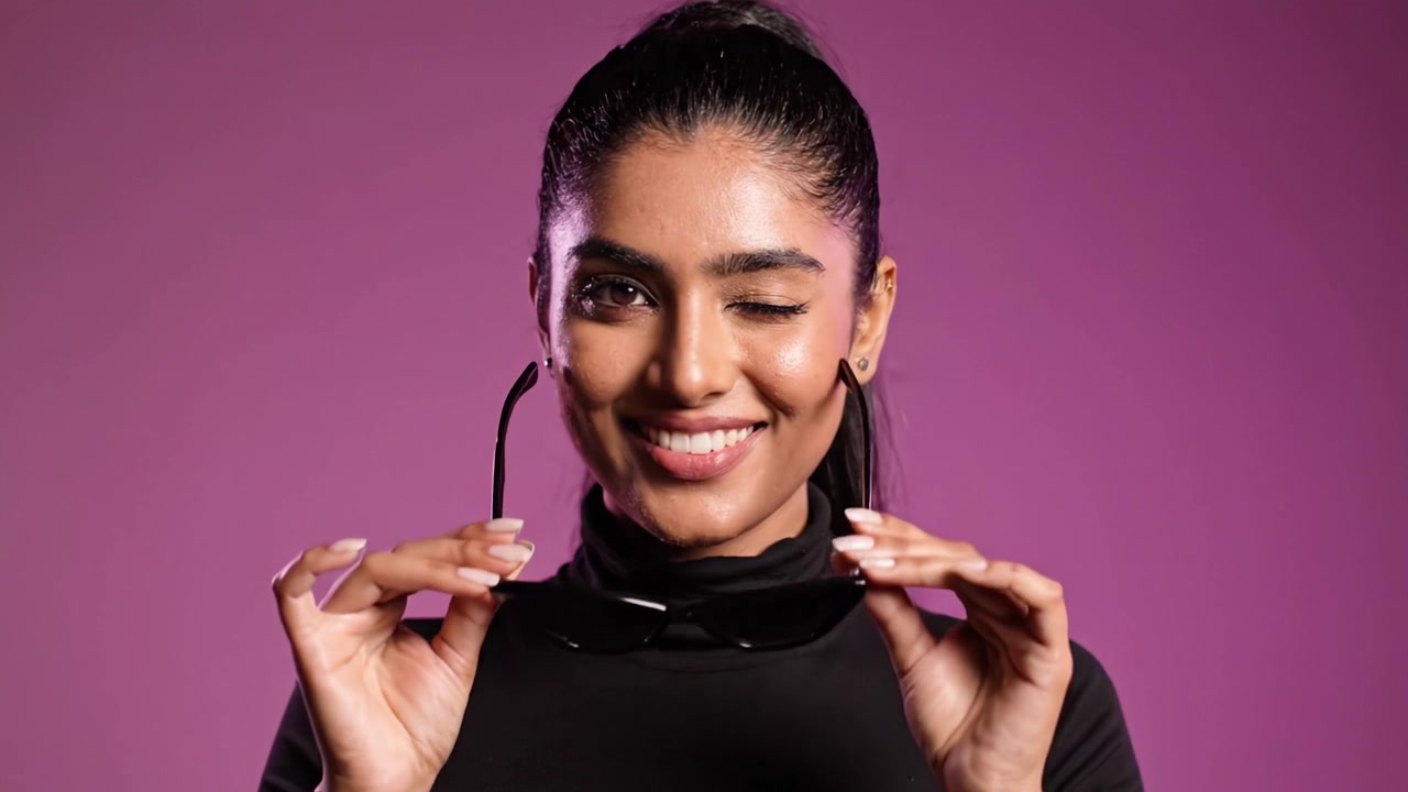 Una giovane donna sicura di sé posa con stile con occhiali da sole su uno sfondo rosa vibrante, mettendo in mostra moda e personalità con eleganza e fascino.