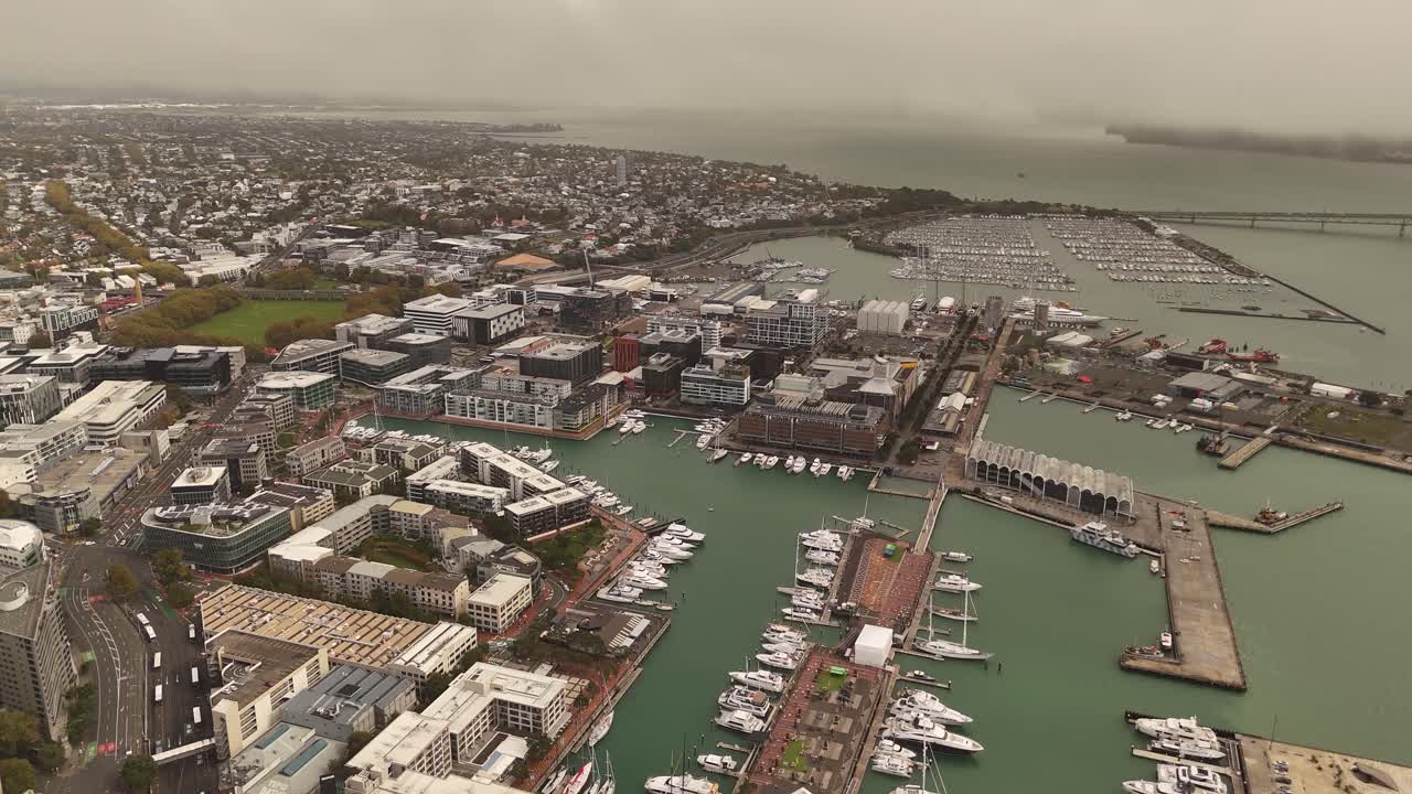 Port and harbor of Auckland City during cloudy day in New Zealand. Aerial wide shot. Coastline of city with parking yachts. Downtown district with homes.