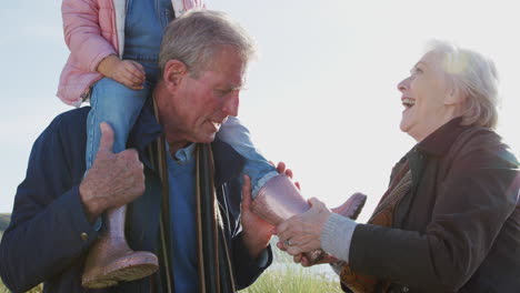 Grandmother-Putting-On-Granddaughters-Wellies-As-Grandfather-Gives-Her-Ride-On-Shoulders