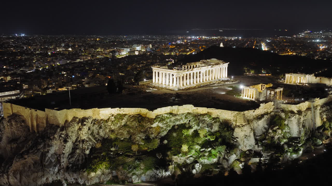 Vista aérea noturna próxima do platô da Acrópole, com o Partenon iluminado em contraste com a Atenas ao redor, enquanto o drone ascende para uma visão panorâmica.