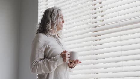 Senior-caucasian-woman-looking-through-window-and-drinking-tea,-slow-motion