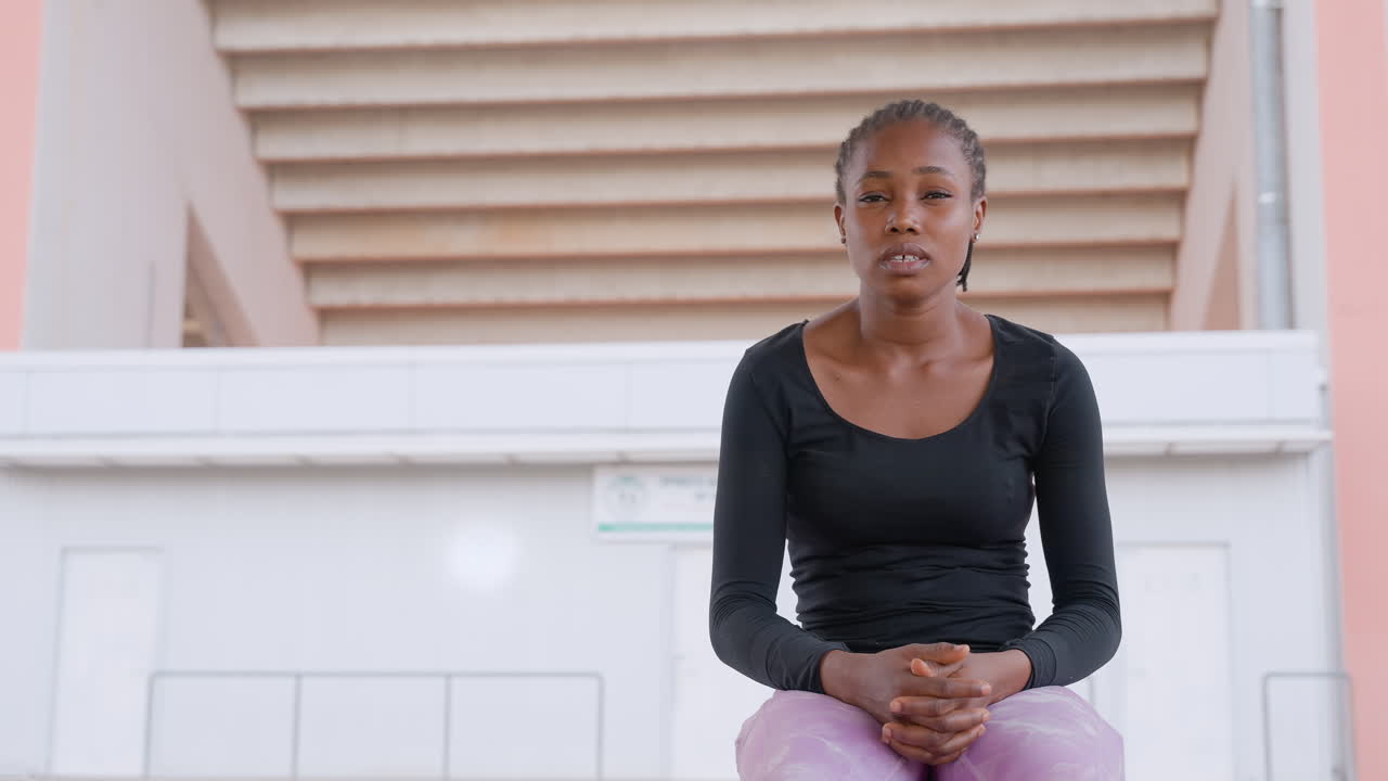 black woman seated on pastel stairs, contemporary dancer pausing between rehearsals with hands clasped and focused expression, soft daylight highlighting warm skin tones and textured braids, minimal