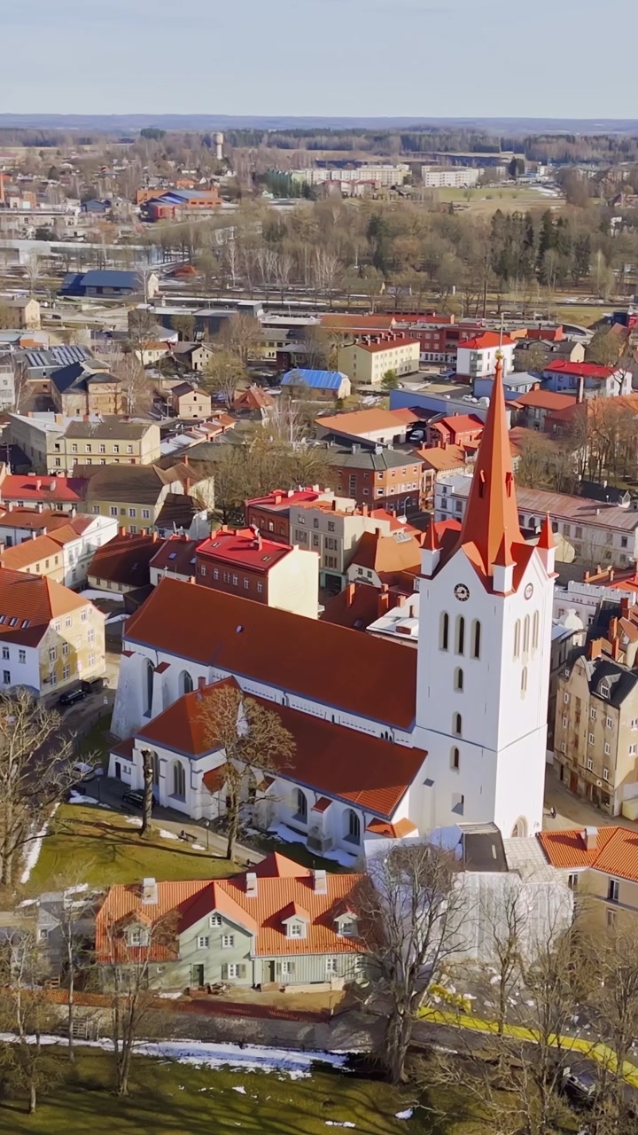 Vertical aerial of a white church with a tall red spire rising above a compact town center, capturing faith, historic architecture, and urban skyline from above.