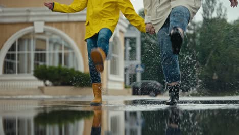 Ein-Glückliches-Mädchen-In-Einer-Gelben-Jacke-Und-Gummistiefeln-Geht-Mit-Ihrer-Mutter-Durch-Eine-Pfütze-Und-Spritzt-Nach-Dem-Regen-Wasser-An-Die-Seiten-Im-Park