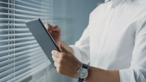 close up view of arabian businessman standing near the window in his office and taping on the tablet, then looking out the window