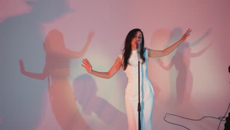 a vocalist wearing a white gown sings passionately while moving her hands,shadows on the wall against a colorful background. she stands in front of a vintage microphone