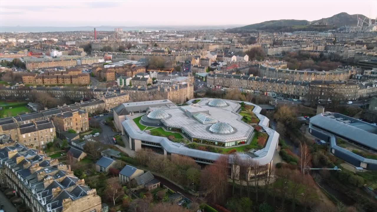 Aerial footage of a modern building in Edinburgh with a unique rooftop design, surrounded by historic architecture and residential areas. The cityscape and distant hills are visible.