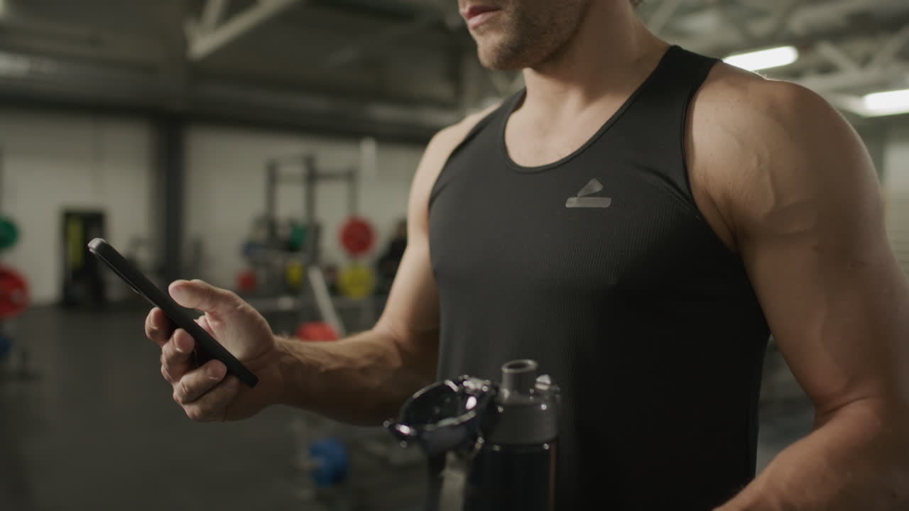 Close-Up of Muscular Man Holding Water Bottle and Using Smartphone in Gym