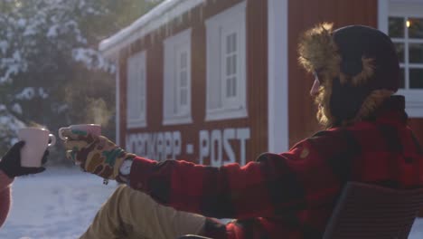 young man in winter clothes holding a hot mug of coffee while sitting on a snowed forest