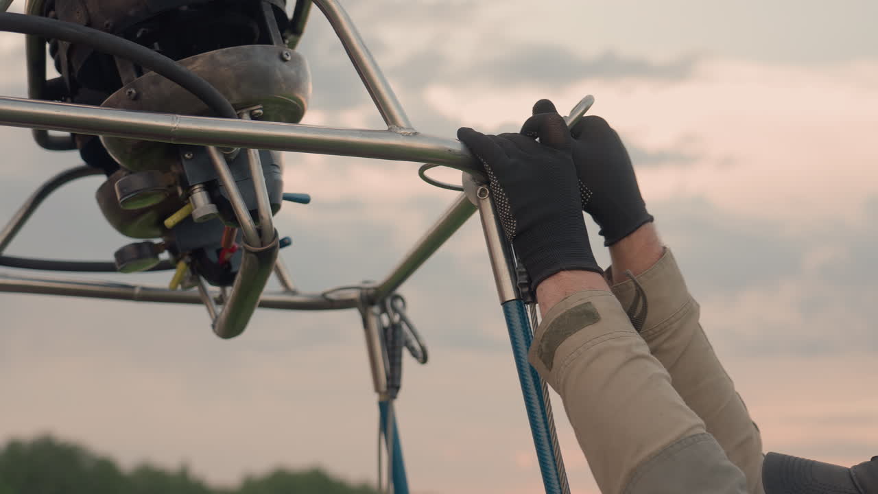 close up of person wearing cap and gloves securing metal hook onto hot air balloon frame with visible ropes hoses and connection points under soft sunset sky during preflight assembly