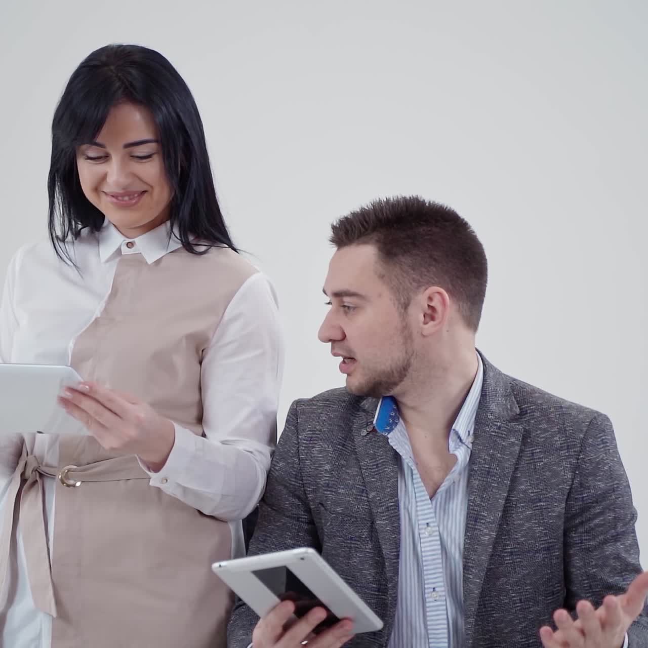 Man and woman are holding gadgets. Handsome man talking to a pretty woman while looking into the tablet isolated in studio. Beautiful lady smiling. Slow motion.