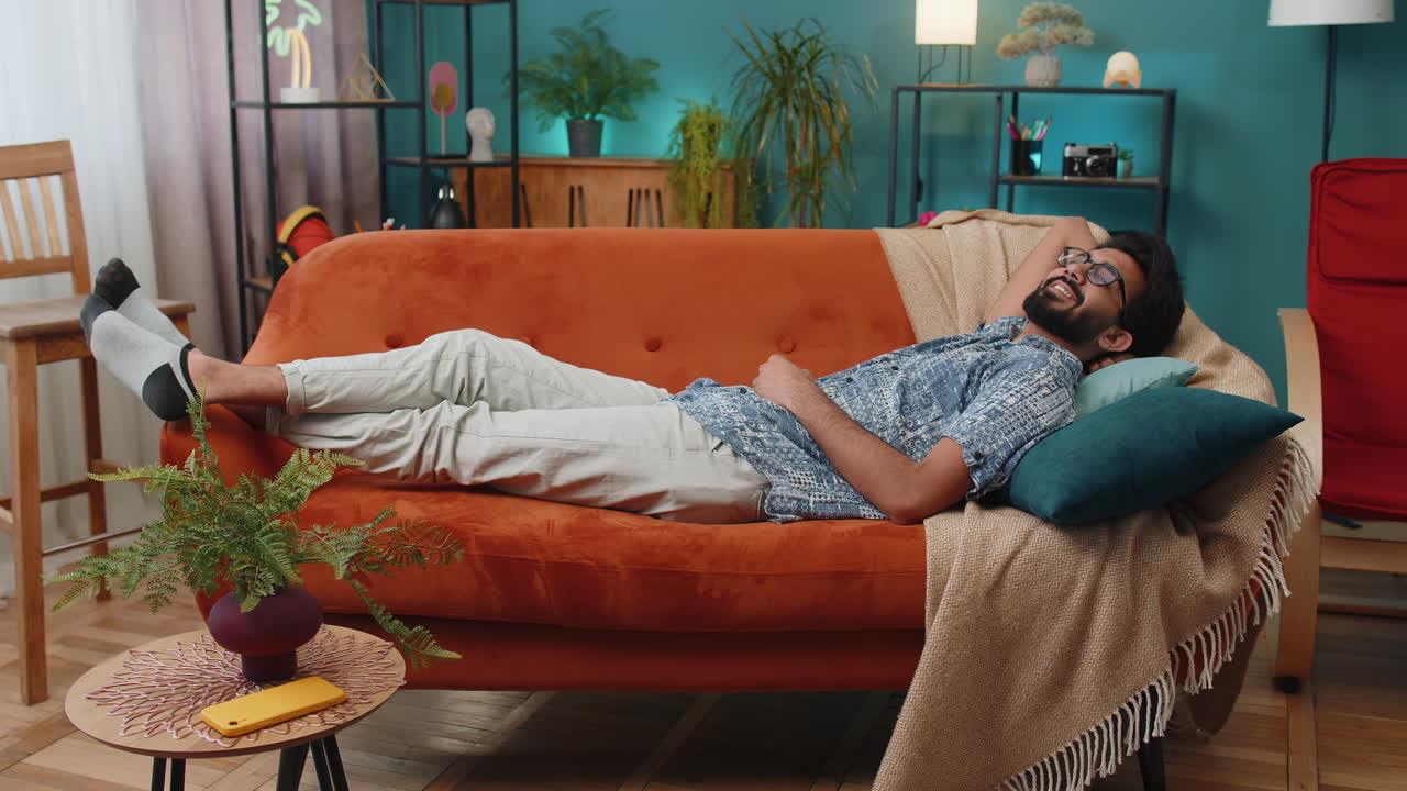 Tired indian man lying down on comfortable couch taking a rest daytime nap relaxing falling asleep