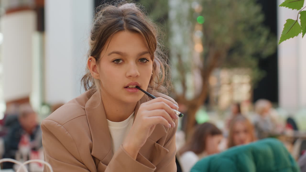 코카서스 십 대 소녀가 카페에서 전자 담배를 피우는 모습, 부드러운 보케와 자연광, 캐주얼 재킷을 입고 붐비는 야외 테라스에서 생각에 잠긴 클로즈업 인물 사진, 표정이 풍부한 얼굴 제스처, 휴대용 전자 담배 기기, 구경하는 사람들