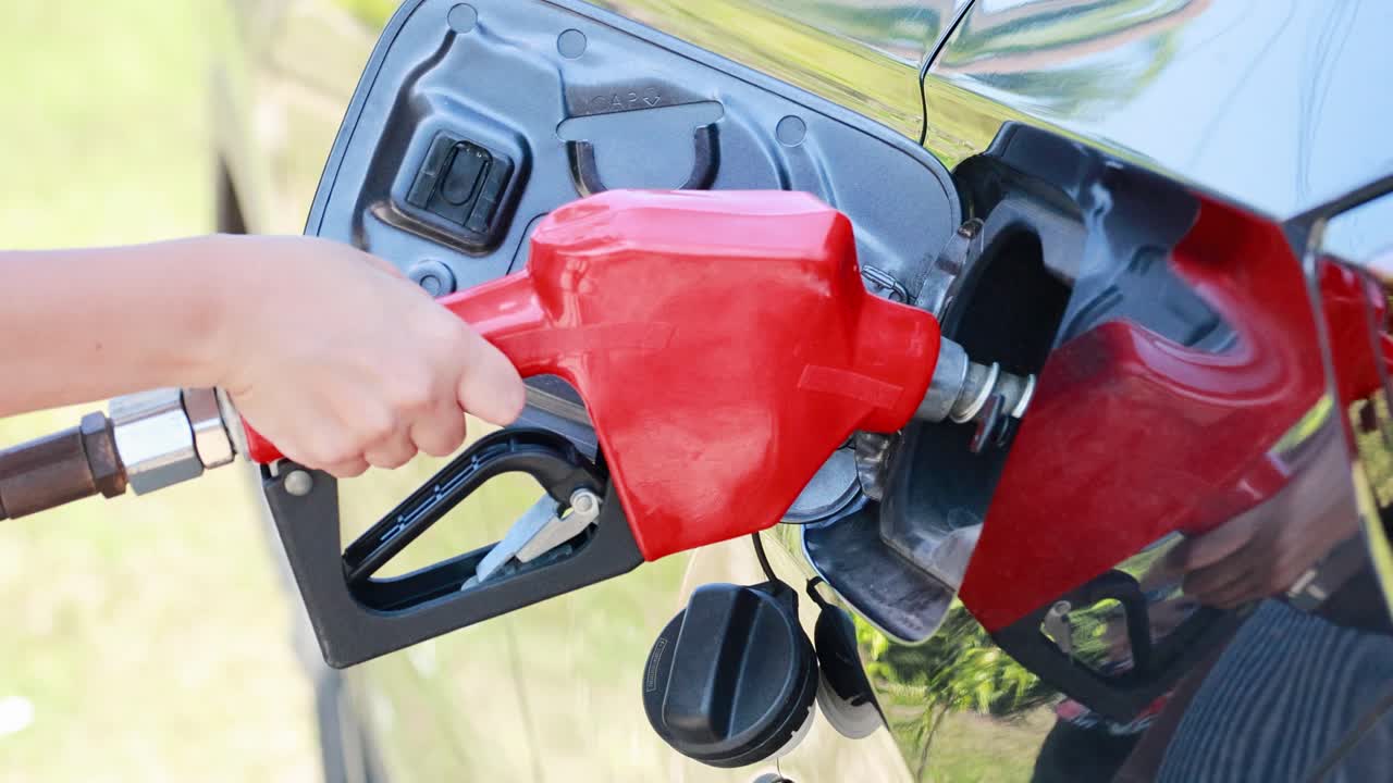 A person refuels a vehicle at a gas station using a red fuel nozzle in daylight