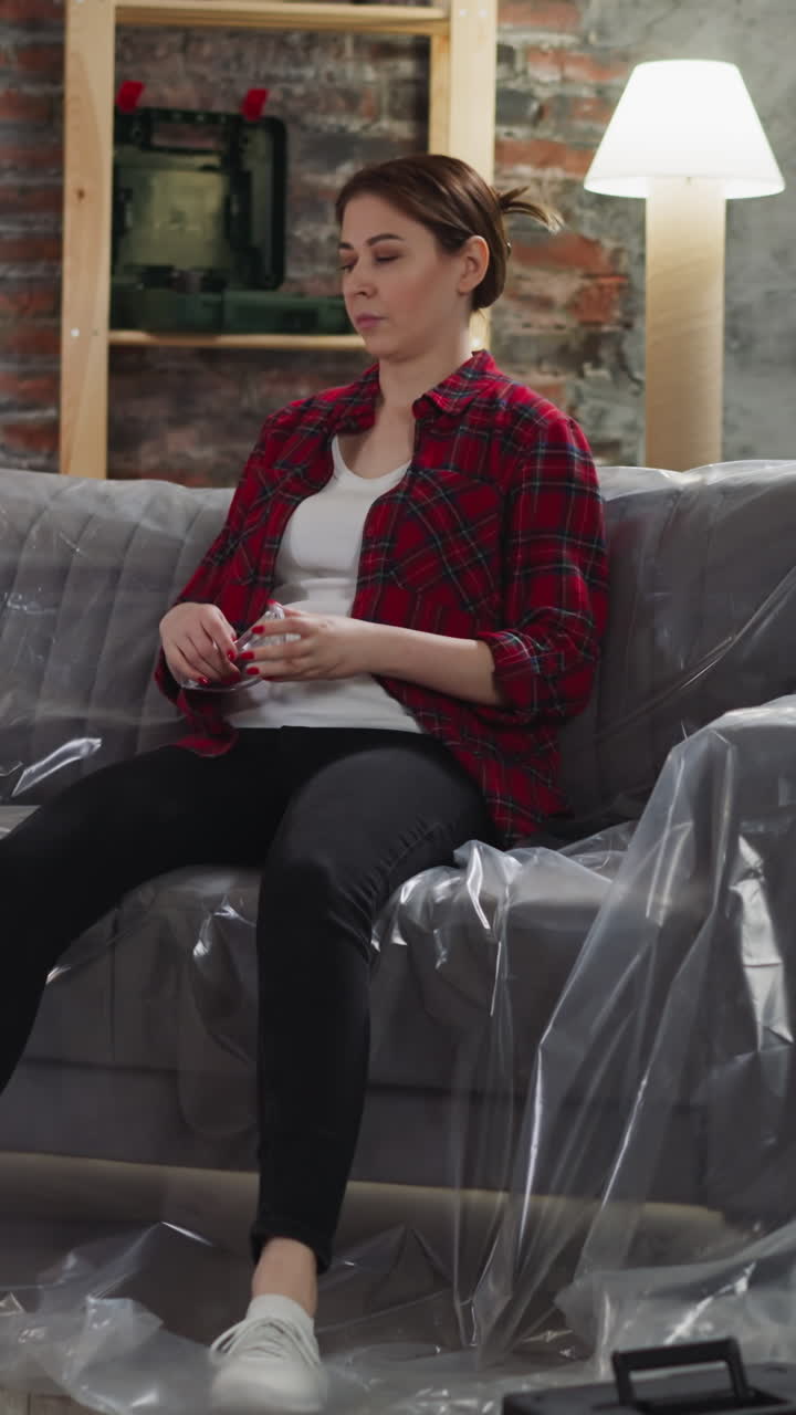 Tired housewife with goggles and drill falls down onto sofa with foil at home. Exhausting repair process in apartment. Handy-woman rests after hard work