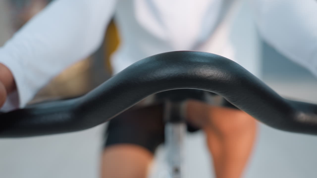 Close up of athlete gripping stationary bike handlebar during intense indoor cycling session in modern gym with blurred exercisers and equipment in background