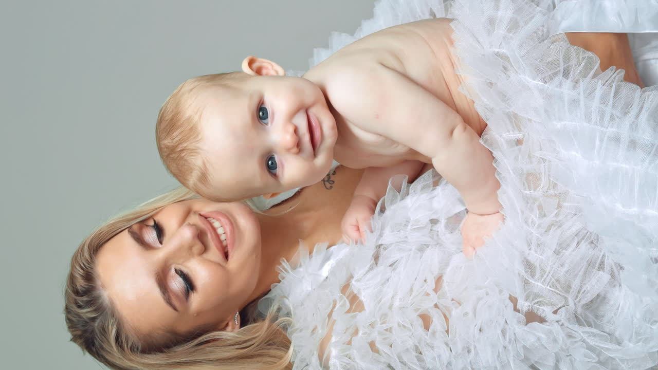 Portrait of a beautiful Caucasian woman with baby in hands. Adorable grey-eyed child looking at camera. White backdrop. Vertical screen.