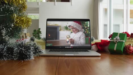 Caucasian-boy-with-santa-hat-playing-with-snow-globe-on-christmas-video-call-on-laptop