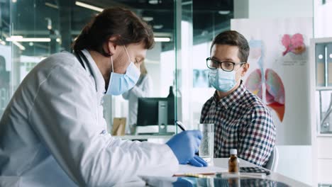 close-up view of male doctor wearing medical mask sitting at desk and speaking with young patient while prescribing a treatment