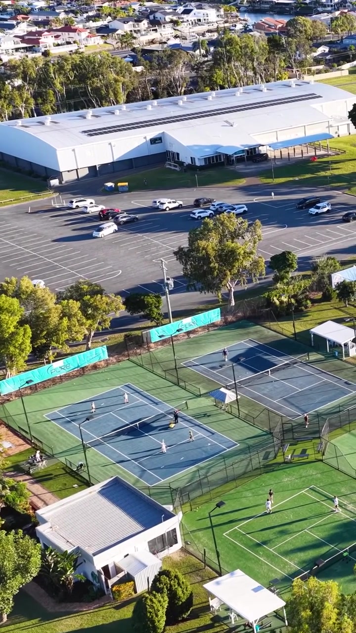 Imágenes de dron mostrando canchas de tenis al aire libre, un estacionamiento y una gran instalación deportiva