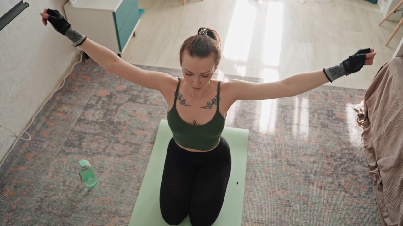 Mulher branca se exercitando em um tapete na sala de estar, vestindo top verde e luvas sem dedos, realizando elevações de braço e alongamentos, com luz solar no tapete, garrafa de água por perto, cenário de quarto aconchegante, calma e focada.