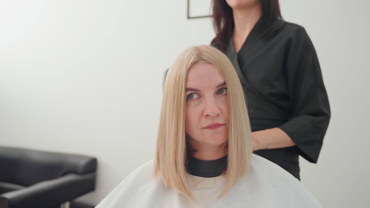 consulenza in un salone di bellezza per una donna bionda sorridente mentre la stilista sistema il caschetto, la ripresa frontale cattura l'espressione, sfondo neutro da studio, atmosfera rilassata e sicura, cliente seduta con mantellina